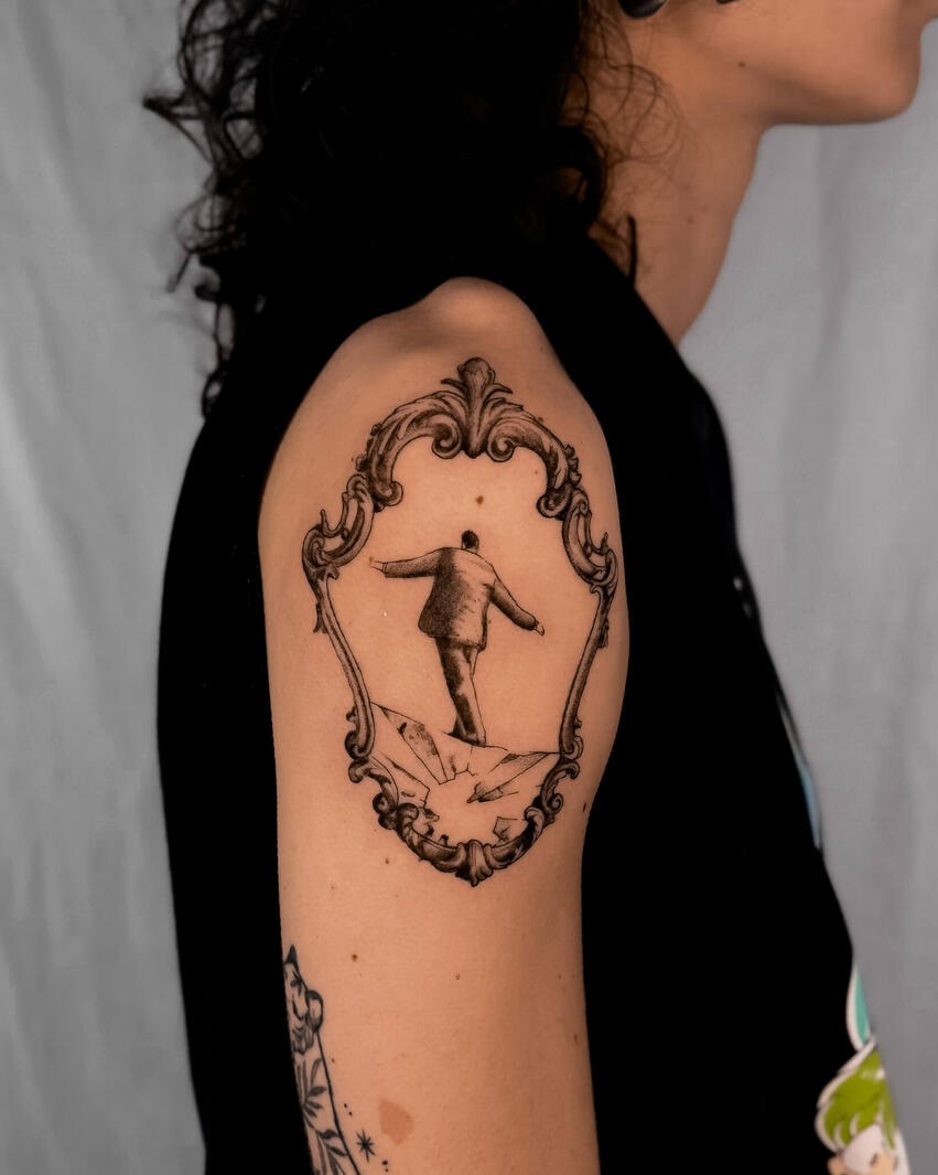 Figura equilibrista nel quadro vintage Tatuaggio fine line di una figura in equilibrio su un vetro all’interno di una cornice vintage ornamentale, con dettagli realistici e ombreggiature soft sul braccio
